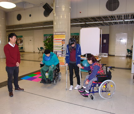This was a game booth for participants to experience using assisting tools to walk like a physically disabled person. The tool the girl in the photo was using is a pair of cane and there was a woman with a wheelchair teaching her. 那裏有遊戲攤位供參加者使用協助工具體驗肢體殘障人士行走。圖中少女使用的工具是一對拐杖，而一位坐着輪椅的女士正在教她使用。