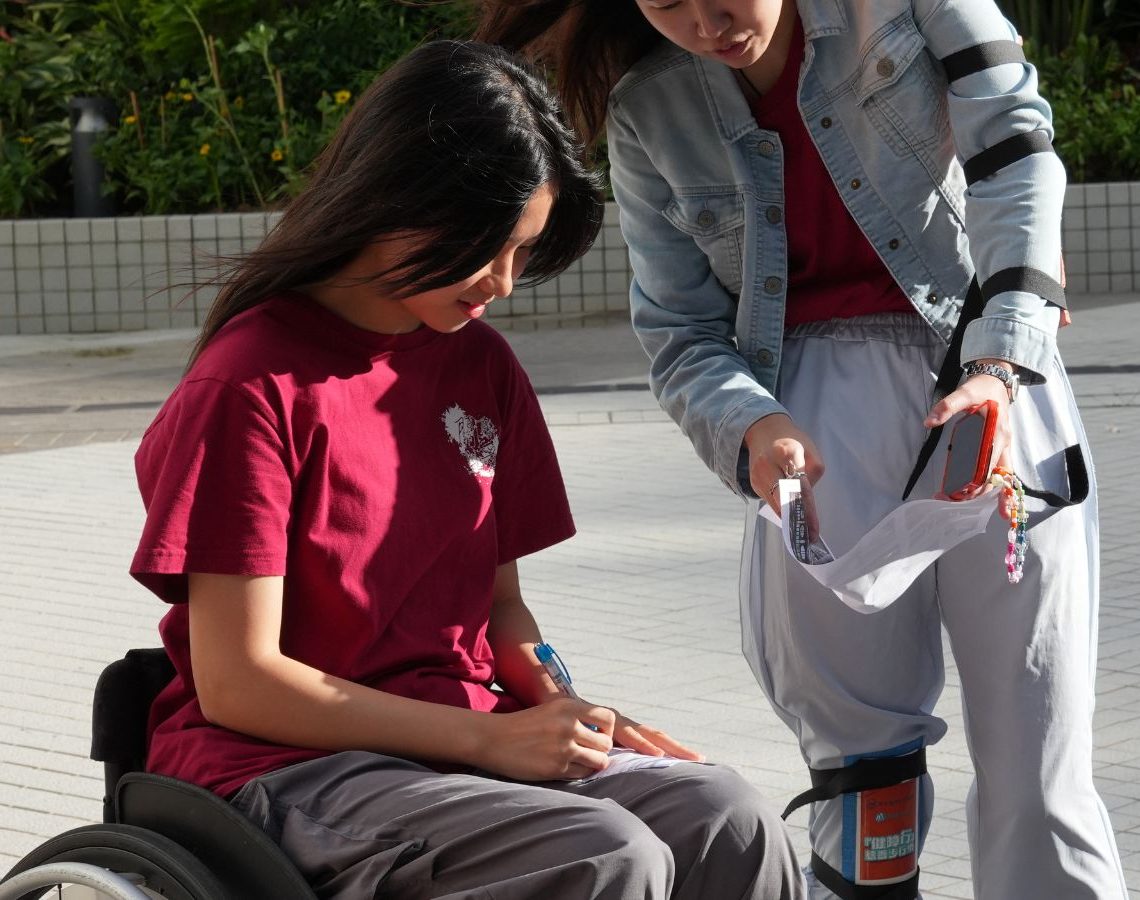 A student seated in a wheelchair fills in a form resting on their lap while a fellow student leans in alongside to help, on a paved campus terrace with planting behind them.