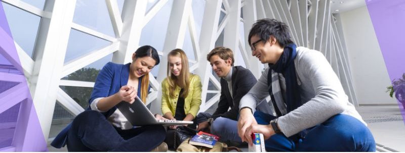 Four students sit together on a campus walkway studying with a laptop and textbooks