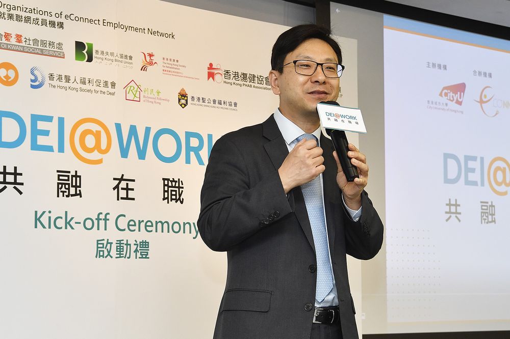 A speaker addresses the DEI@WORK kick-off ceremony beside a backdrop listing the eConnect Employment Network member organisations