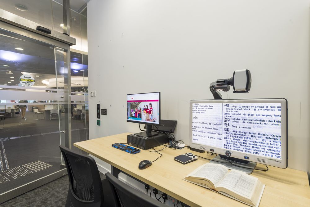 A study workstation in the library Special Education Room, equipped with a video magnifier displaying an enlarged dictionary page and an accessible large-key keyboard