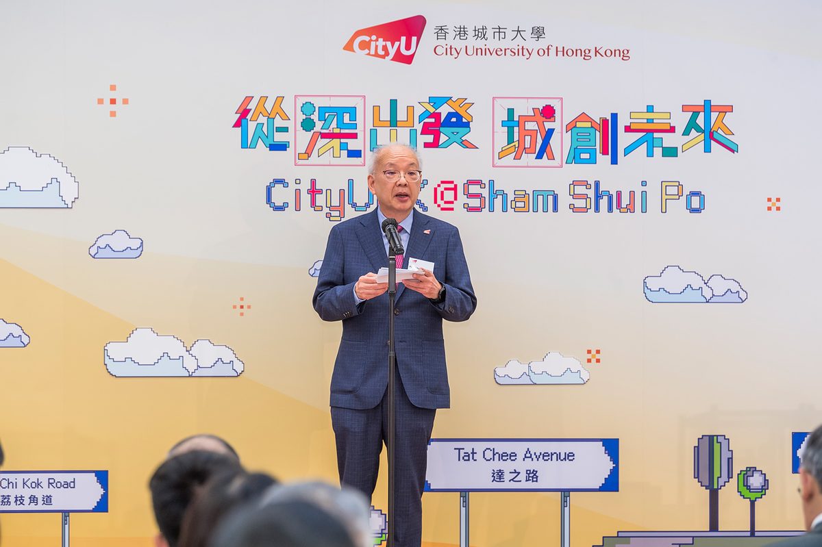 A speaker addressing guests from a microphone in front of the "CityUHK@Sham Shui Po" exhibition backdrop, a pixel-art Hong Kong skyline with Tat Chee Avenue and Lai Chi Kok Road street signs