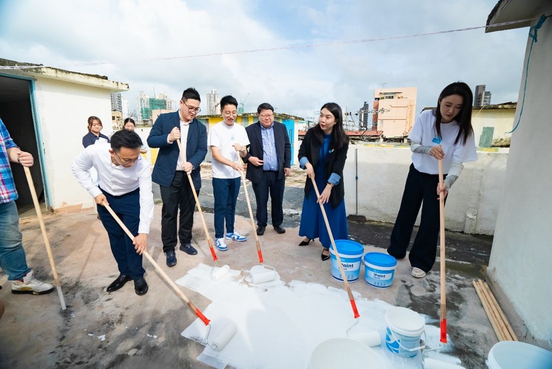 Guests and CityUHK staff use long-handled rollers to spread white cooling paint across a Sham Shui Po rooftop at the project launch, with tubs of iPaint beside them and the surrounding tenement blocks behind.